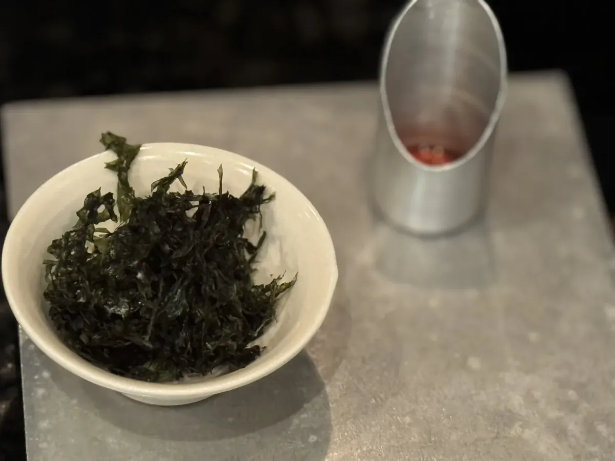 A small white bowl holding dark green steeped tea leaves, on a pale stone counter
