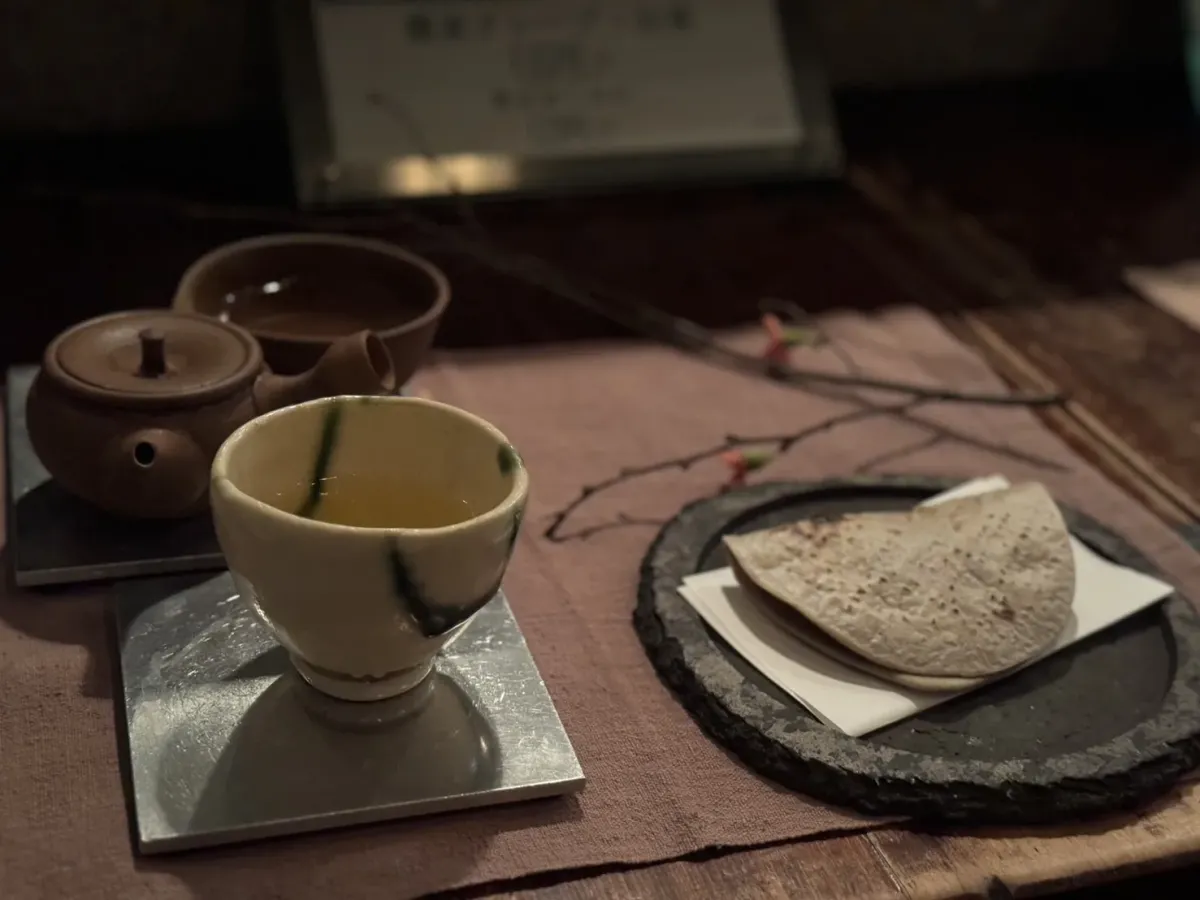 A cup of pale green tea on a metal plate beside a small clay kyusu and a dry sweet on a dark slate dish, with a branch arrangement behind