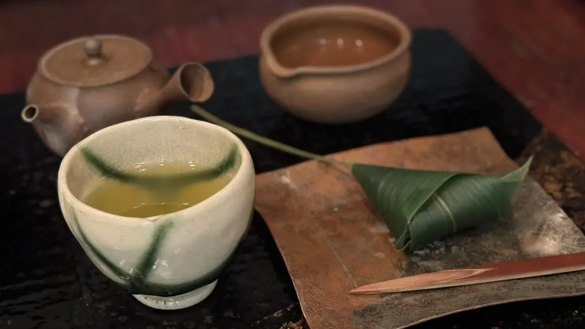 A cream and green tea bowl of pale sencha beside a small clay kyusu and a sweet wrapped in a bamboo leaf, on a dark counter