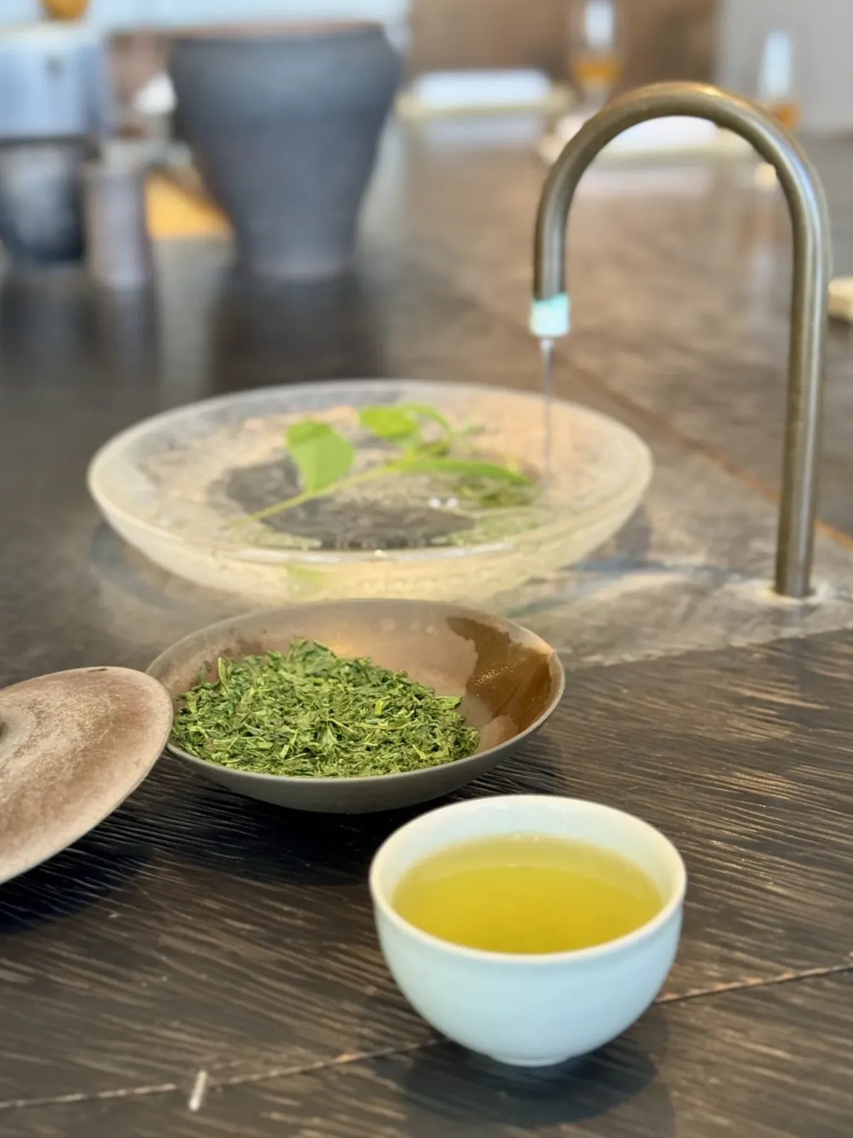 A slim brass gooseneck tap at the counter running water into a glass dish, beside a shallow bowl of dry green tea leaves and a cup of tea