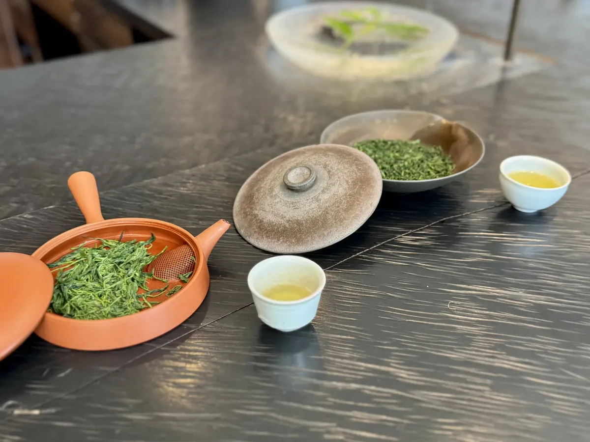 A dark wood counter with a vermilion roasting vessel holding fresh green tea leaves, a lidded pot, a shallow dish of dry leaves, and two small white cups of pale green tea