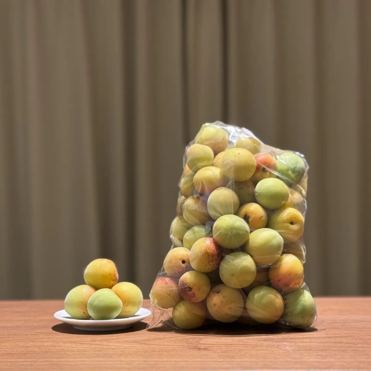 Hard green ume plums in a net bag, with a few loose plums on a small white dish